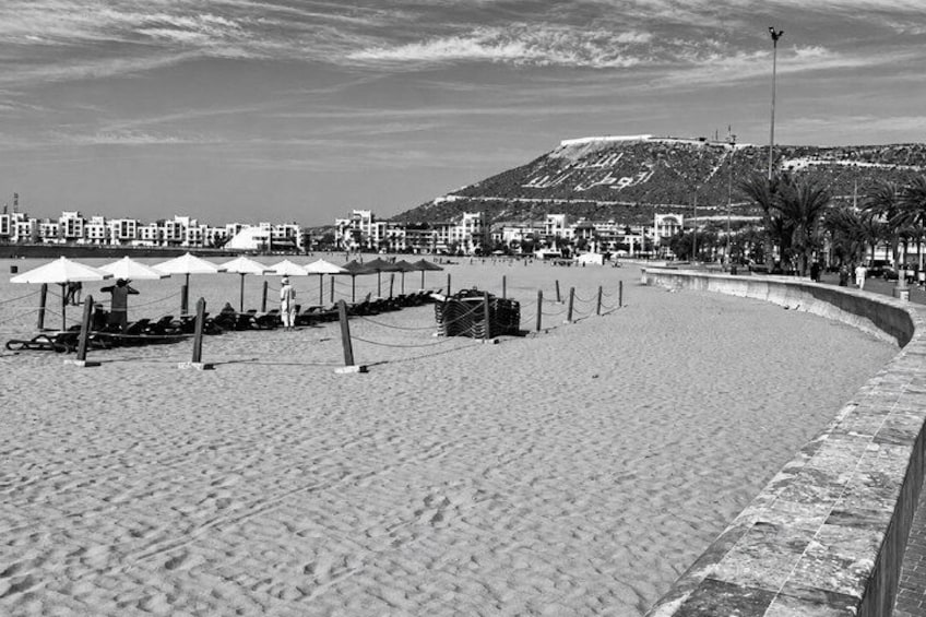 Una playa con sombrillas y una cerca, una colina al fondo y edificios a lo largo de la costa. 4