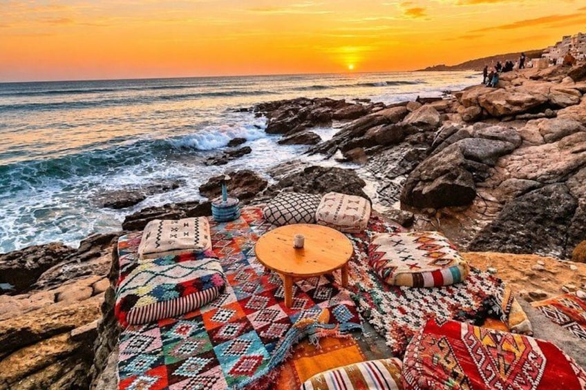 Un atardecer costero con una playa rocosa, esteras tejidas de colores y una pequeña mesa redonda. 2