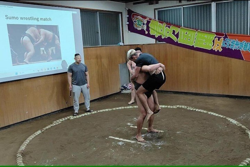 Un combate de sumo en curso, con un proyector mostrando una imagen relacionada al fondo. 4