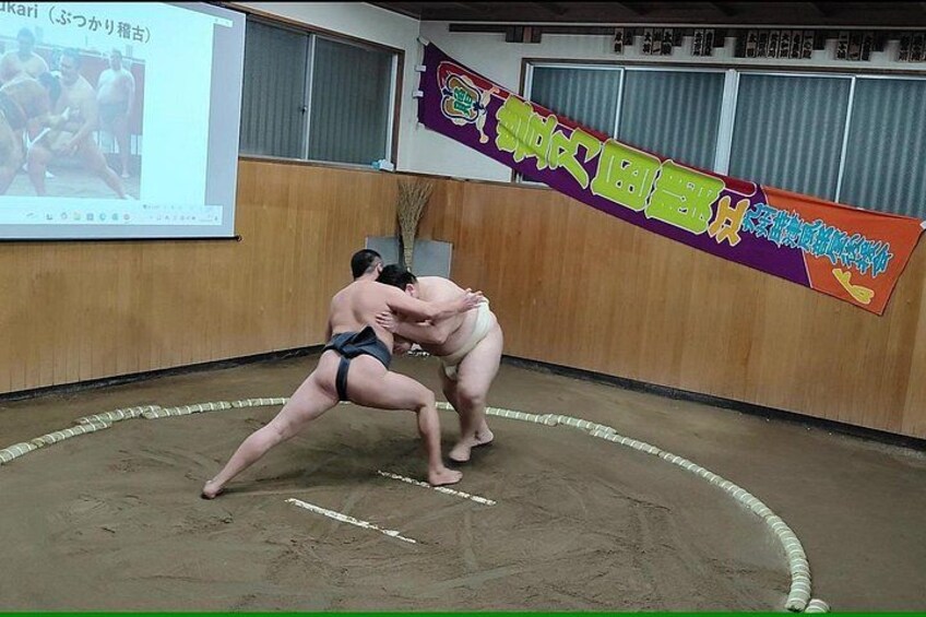Dos luchadores de sumo en un ring, una pantalla de proyector y un cartel con texto en japonés. 4