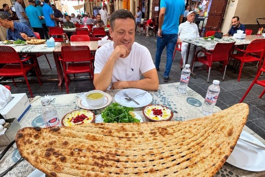 A man sitting at a table with a large, grilled flatbread, salad, and water bottles. 5