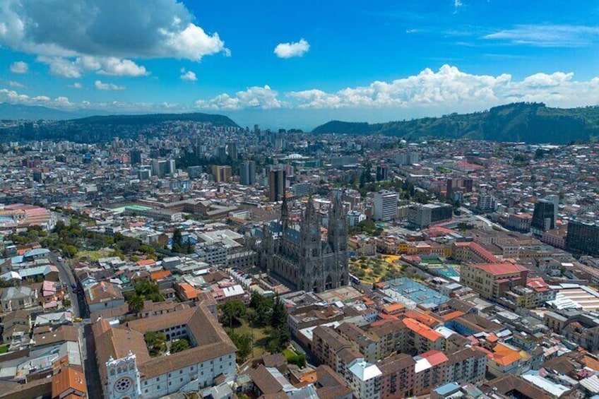 An aerial view of a city with a prominent cathedral, dense urban buildings, and a distant mountain range under a clear sky. 2