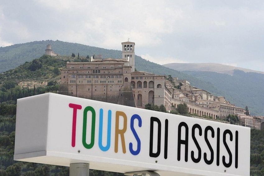 En skylt med texten ”TOURS DI ASSISI” i färgglada bokstäver, en historisk byggnad med valv och ett bergigt landskap i bakgrunden. 1