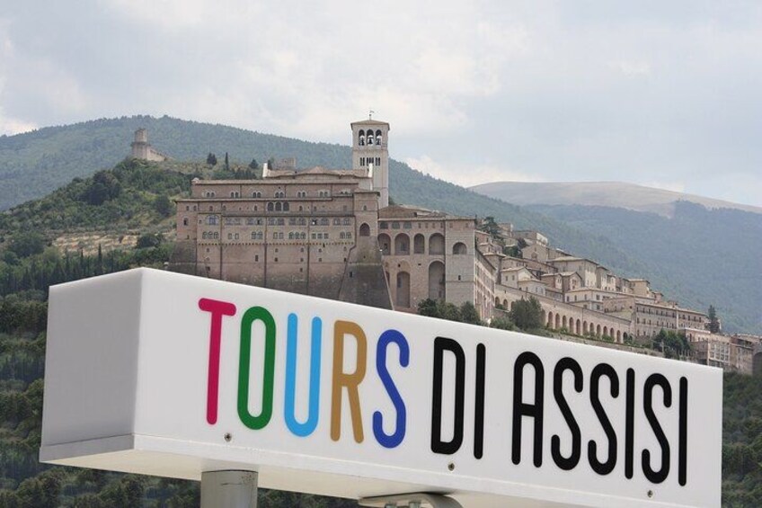 Skilt med 'TOURS DI ASSISI' i farverige bogstaver, en historisk bygning og et bjergrigt landskab i baggrunden. 1