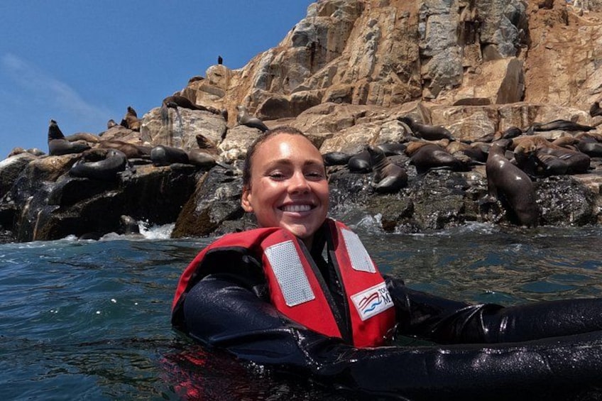 Una persona con chaleco salvavidas rojo flotando en el agua, con acantilados rocosos y lobos marinos al fondo. 3