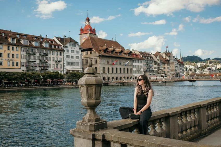 A woman sitting on a bridge railing with a river and buildings in the background. 1