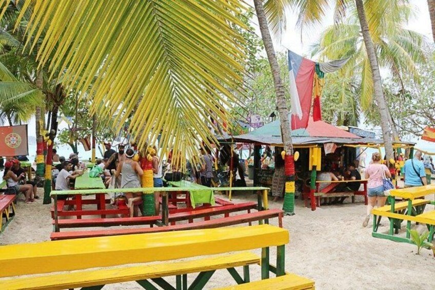 A beachside gathering with people seated at colorful wooden tables and benches, under palm trees. 2
