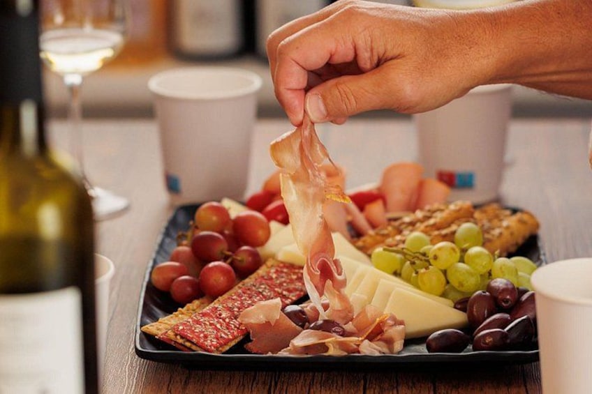 Una persona sostiene un trozo de embutido, junto a una tabla de fiambres con varios quesos, uvas y aceitunas. 2