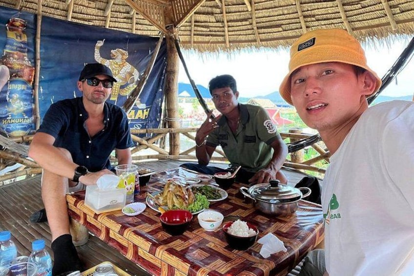 A group of people sitting at a table with food and drinks, under a thatched roof. 2