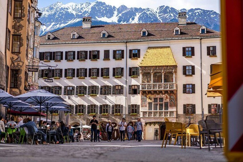 Une rue avec des terrasses, un bâtiment orné d’une fresque murale et, au loin, des montagnes aux sommets enneigés. 2