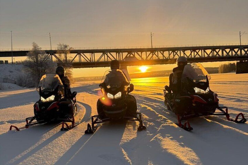 Drei Schneemobile auf einem verschneiten Pfad, im Hintergrund eine Brücke. 3