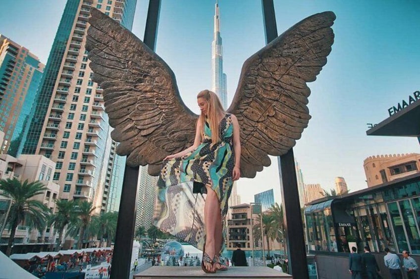 A person in a dress with large wings, standing in front of a building with a sign that reads 'EMAAR'. 4