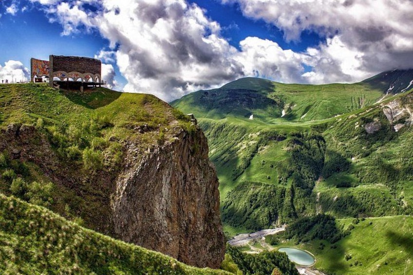 A mountainous landscape with a building on a cliff, lush greenery, and a river. 1