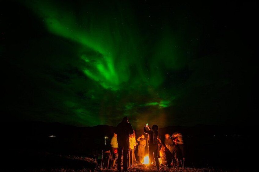 Un groupe de personnes s’est rassemblé autour d’un feu de camp, sous un ciel nocturne illuminé par des aurores boréales vertes. 4
