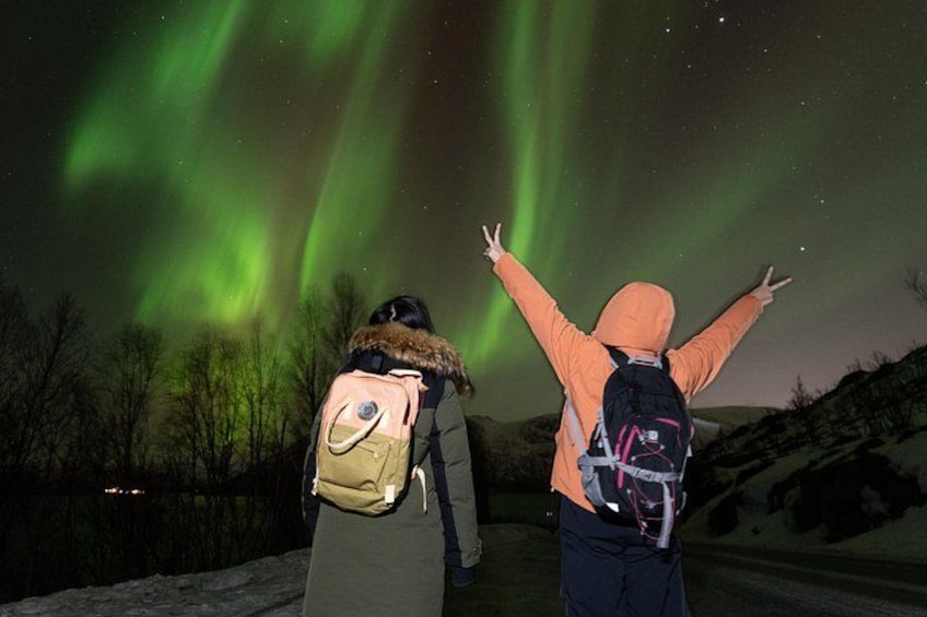 Twee personen buiten, waarvan er één de armen omhoog heeft, kijkend naar de nachthemel met groene aurora's. 2