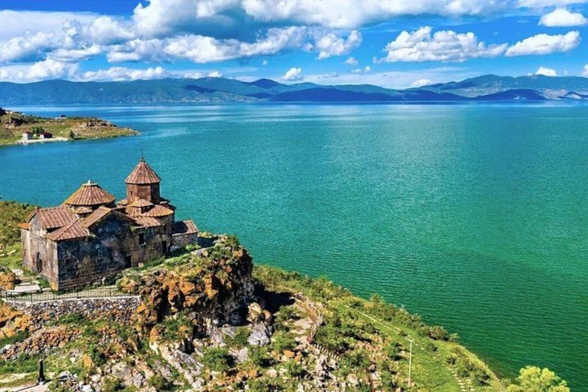 Une église historique, perchée sur une falaise rocheuse, surplombe une vaste étendue d’eau, avec des montagnes en toile de fond. 1