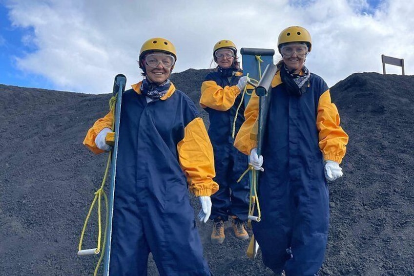 Tre personer med skyddsutrustning, inklusive hjälmar och handskar, som håller i verktyg och står på en hög av mörkt material, troligen kol. I bakgrunden syns en stol och en molnig himmel. 5