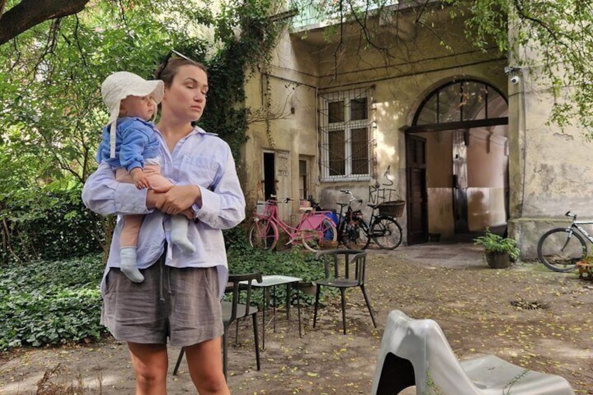 Une personne tenant un enfant dans un jardin, avec des vélos et un bâtiment en arrière-plan. 2