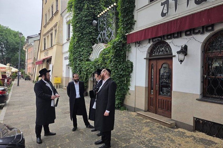 A street scene with buildings, a man in a suit, and a man in a traditional Jewish prayer shawl. 1