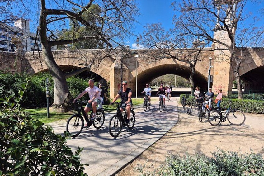 Cyclists on a paved path under a stone bridge with greenery on the sides. 4