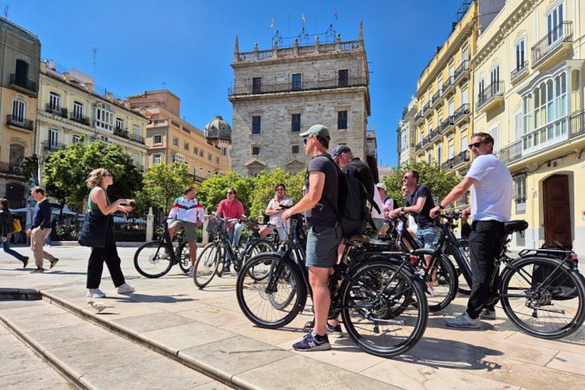 A group of people with bicycles in an urban setting, a historic building in the background, and a clear sky. 2