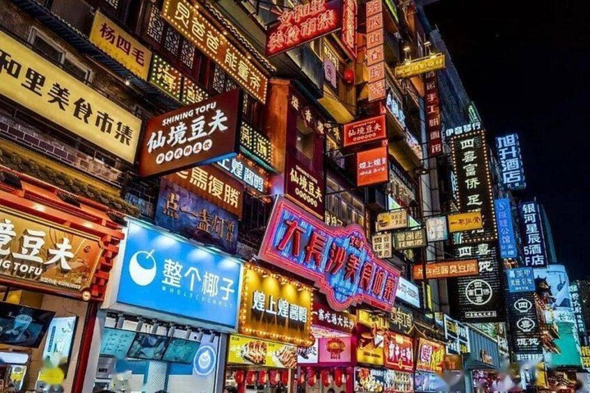 Una calle luminosa de noche con numerosos carteles iluminados en un idioma extranjero, indicando una zona comercial con tiendas y restaurantes. 5