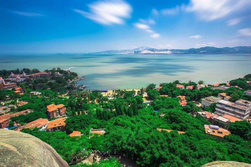 A coastal town with dense greenery, buildings, and a calm sea under a clear sky. 1