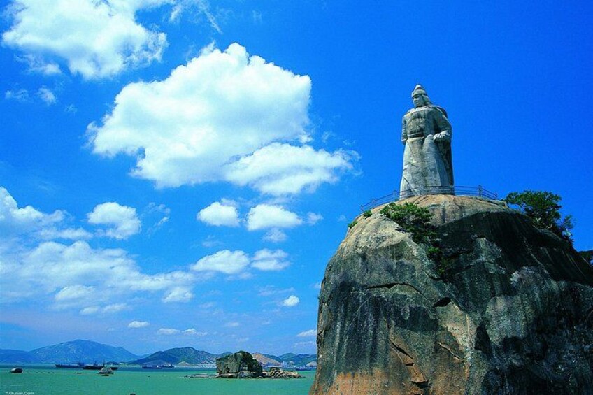 A large rock with a statue on top, a clear blue sky, and calm sea with boats. 5
