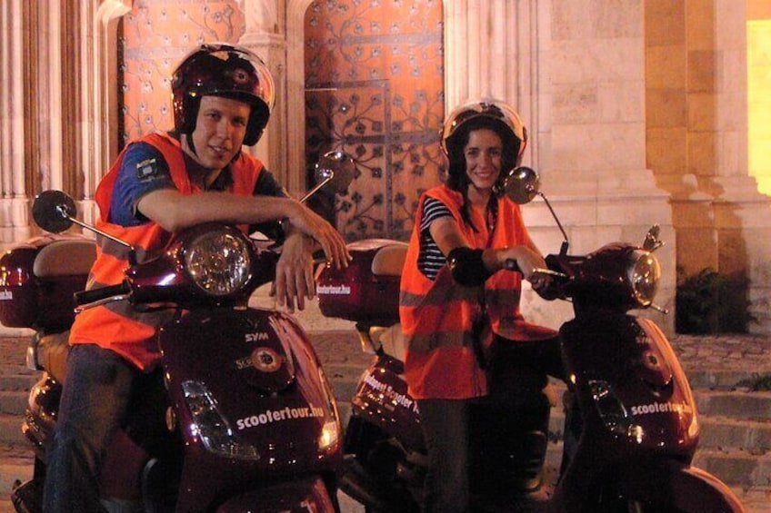 Twee personen op scooters met 'scooterour.no' branding, geparkeerd voor een gebouw met kolommen en een patroon in de muren. 1