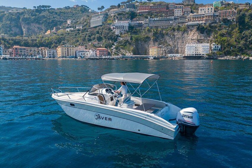 A white motorboat with a canopy, named 'JAVER,' on calm water with a coastal town and cliffs in the background. 4