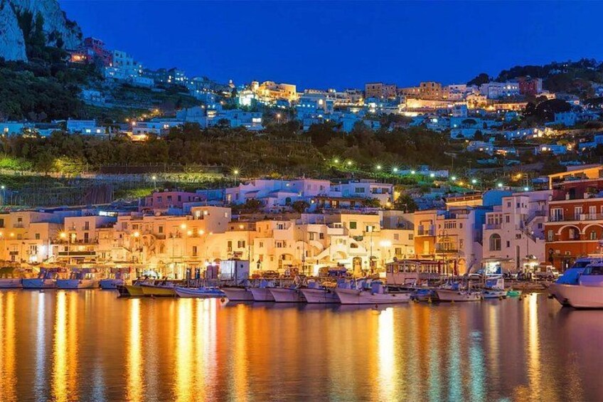 A coastal town at dusk with illuminated buildings and boats docked in the harbor. 1