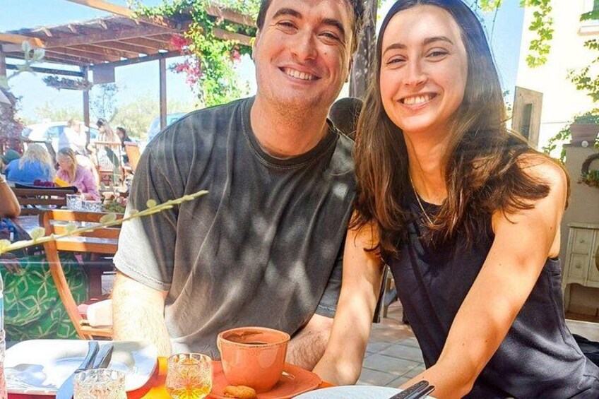 Two people sitting at an outdoor table with a drink and a plate of food. 2