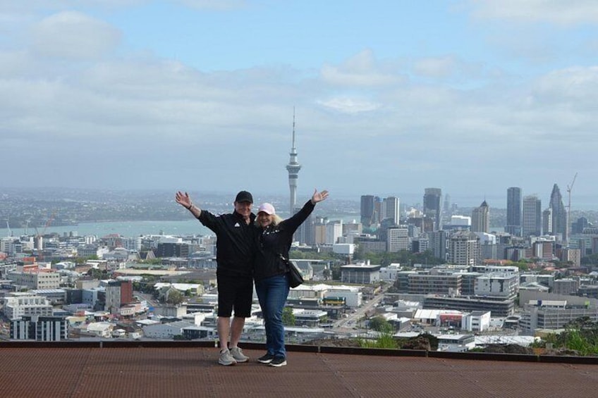Two people standing on a rooftop with arms outstretched, overlooking a cityscape with a prominent tower. 1