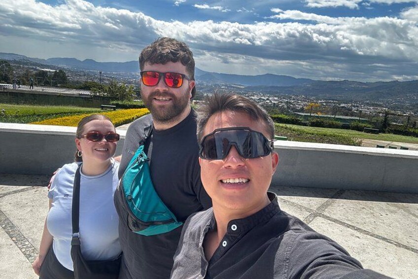 Tre personer som tar en selfie med en vacker utsikt över berg och en stadsbild i bakgrunden. 1