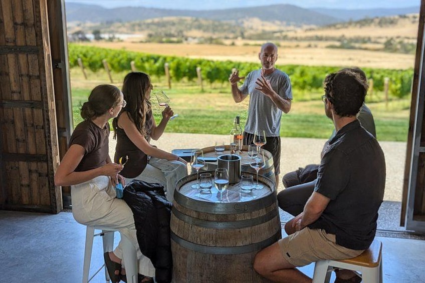 A group of people sitting around a barrel table outdoors with a vineyard in the background. 4