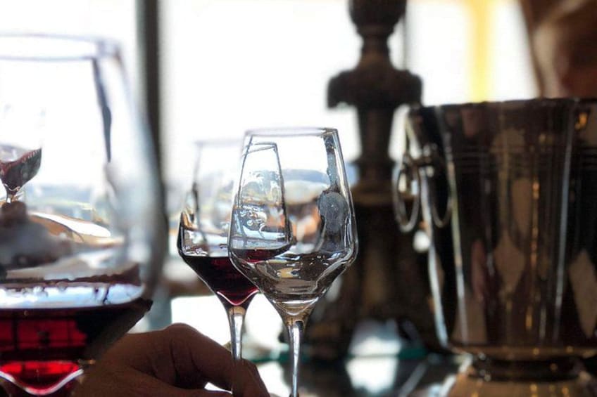 A hand holding a wine glass with a red liquid, a wine bottle, and a metal container in the background. 3