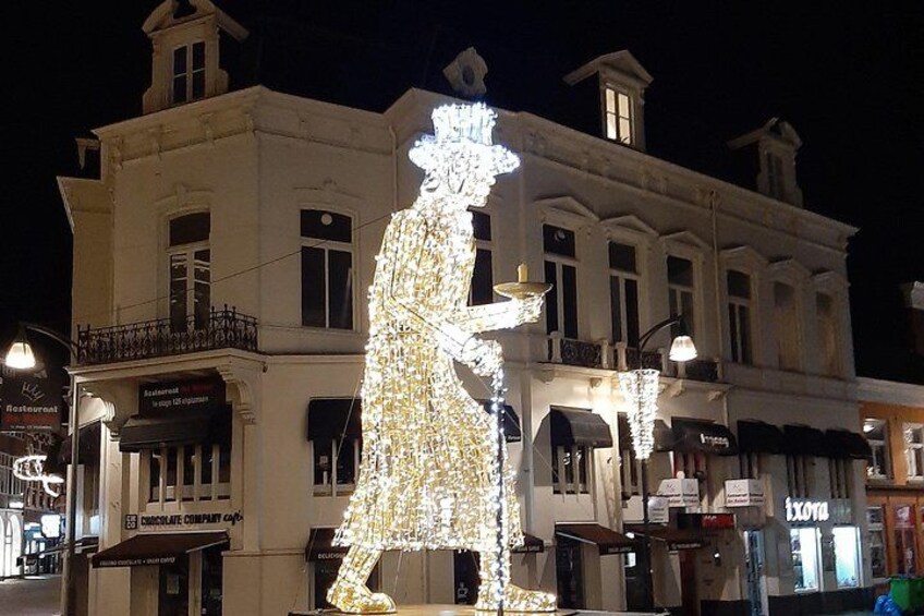 A large illuminated sculpture of a person holding a lighted object, with buildings and shops in the background. 4