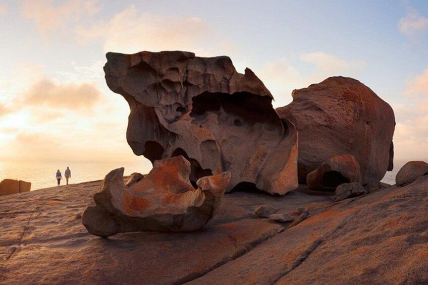 A large rock formation with a cave-like opening, two people walking in the background, and a sunset sky. 1