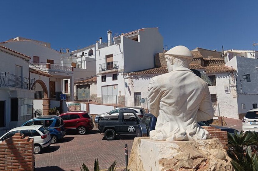 A white statue of a man in a white robe, parked in front of a row of white buildings with red-tiled roofs, under a clear blue sky. 2