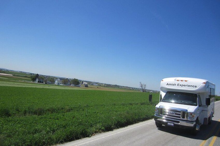 A white RV named 'Amish Experience' driving on a road with green fields and a clear blue sky in the background. 3