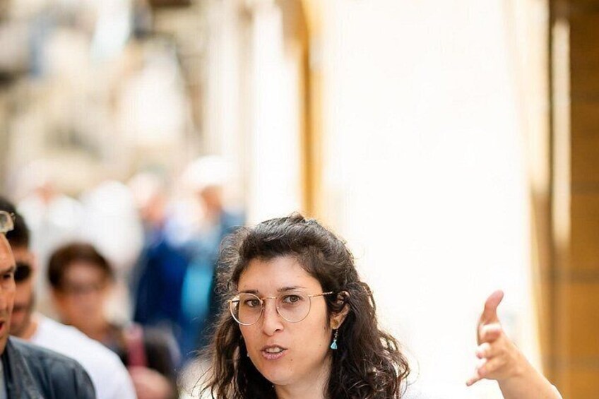 A person gesturing with their hand in an outdoor setting with blurred figures in the background. 4