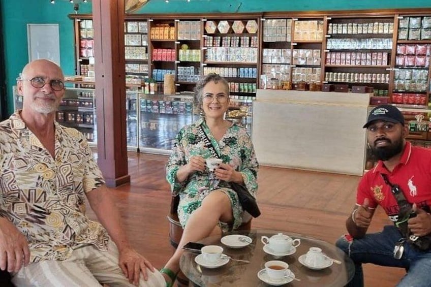 Three people seated at a table with cups and saucers, in a store with shelves of products in the background. 5