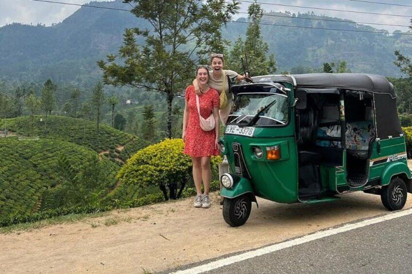 Dos personas de pie junto a un auto-rickshaw verde en una carretera con un paisaje montañoso al fondo. 1