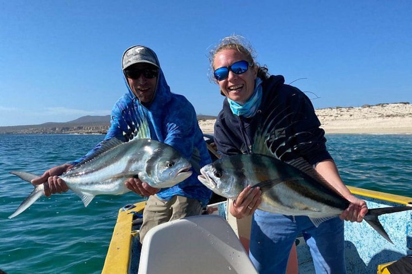 Two people holding large fish on a boat with a beach and water in the background. 3