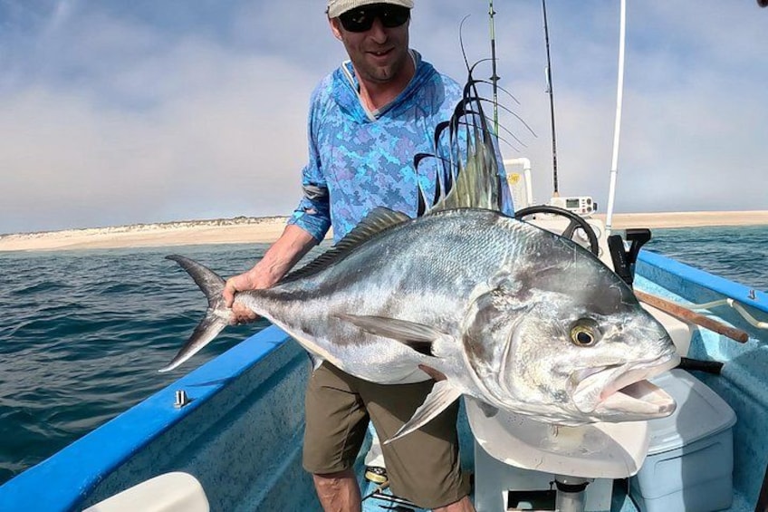 A person holding a large fish on a boat with a sandy beach and calm sea in the background. 5