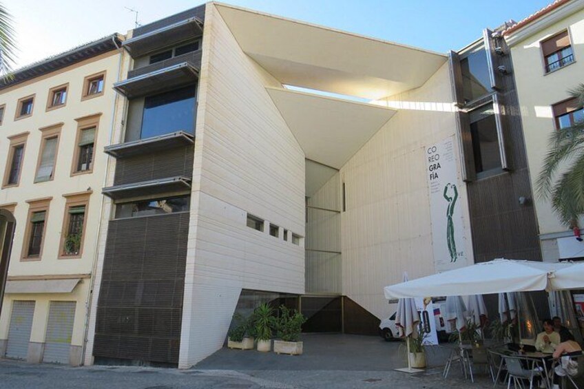 Un bâtiment moderne avec un toit en pente et une grande affiche sur sa façade. On y trouve des tables et des chaises à l’extérieur, ainsi qu’un fourgon blanc garé à proximité. 2