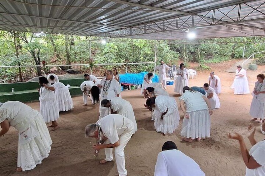 Un grupo de personas vestidas de blanco reunidas al aire libre bajo un techo metálico, algunas arrodilladas y otras de pie, con árboles y un lomo azul al fondo. 2