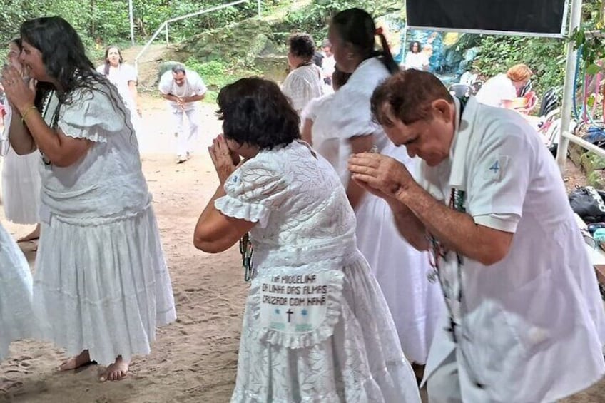 Personas con vestidos blancos reunidas en una playa arenosa, algunas con las manos juntas en oración. 1