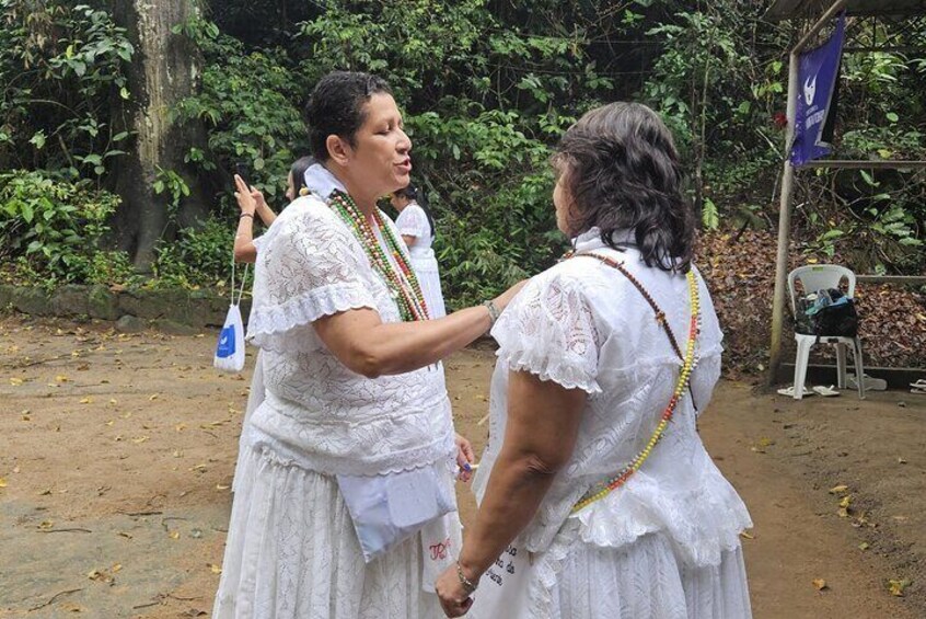 Dos mujeres con vestidos de encaje blanco y collares de cuentas, de pie al aire libre con follaje de fondo. 3