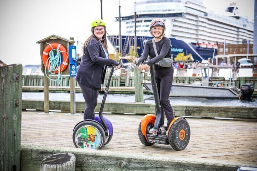 Two people on segways on a wooden boardwalk by a waterfront with a lifebuoy and a building in the background. 2